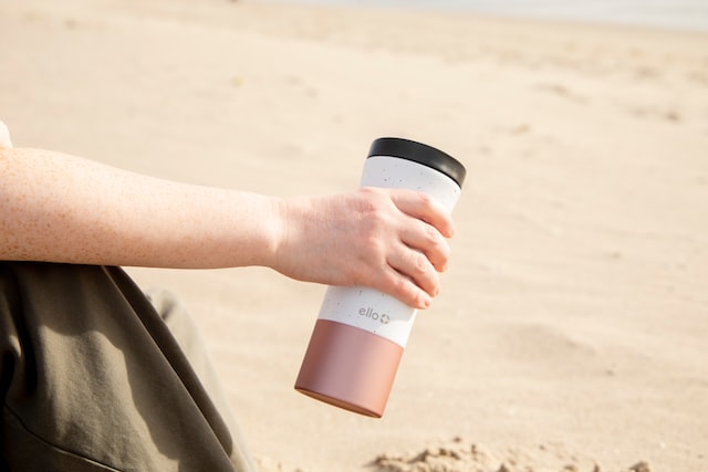person on travel, sitting and holding to-go tumbler cup with sand in background