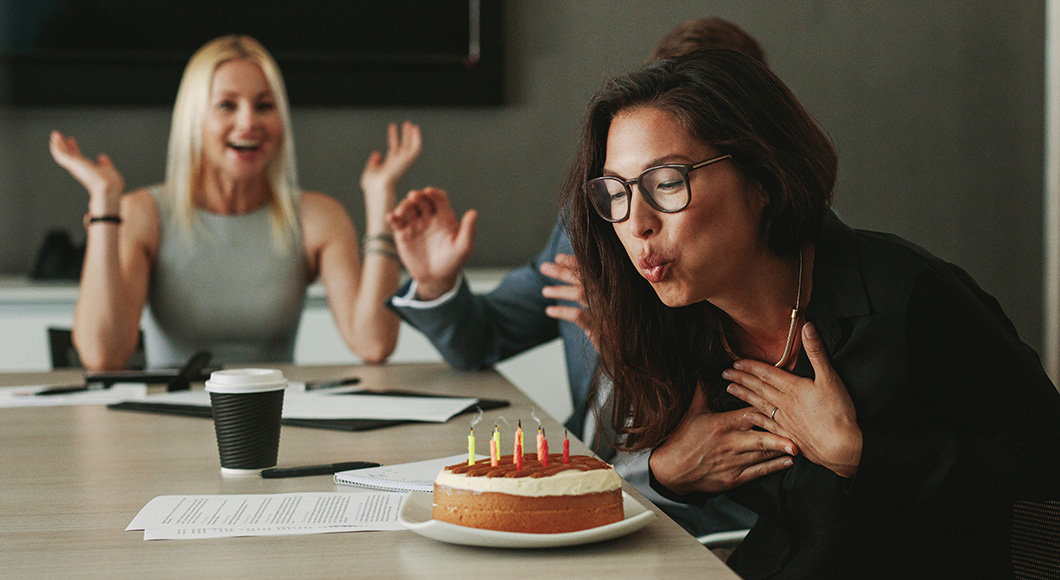 Employees celebrating birthday including employee blowing out candles on cake which is on conference room table with papers, notebook, coffee cup and pens.