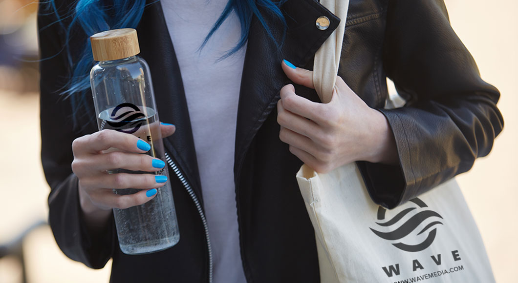 Person holding glass water bottle with bamboo lid, and carrying cotton tote.