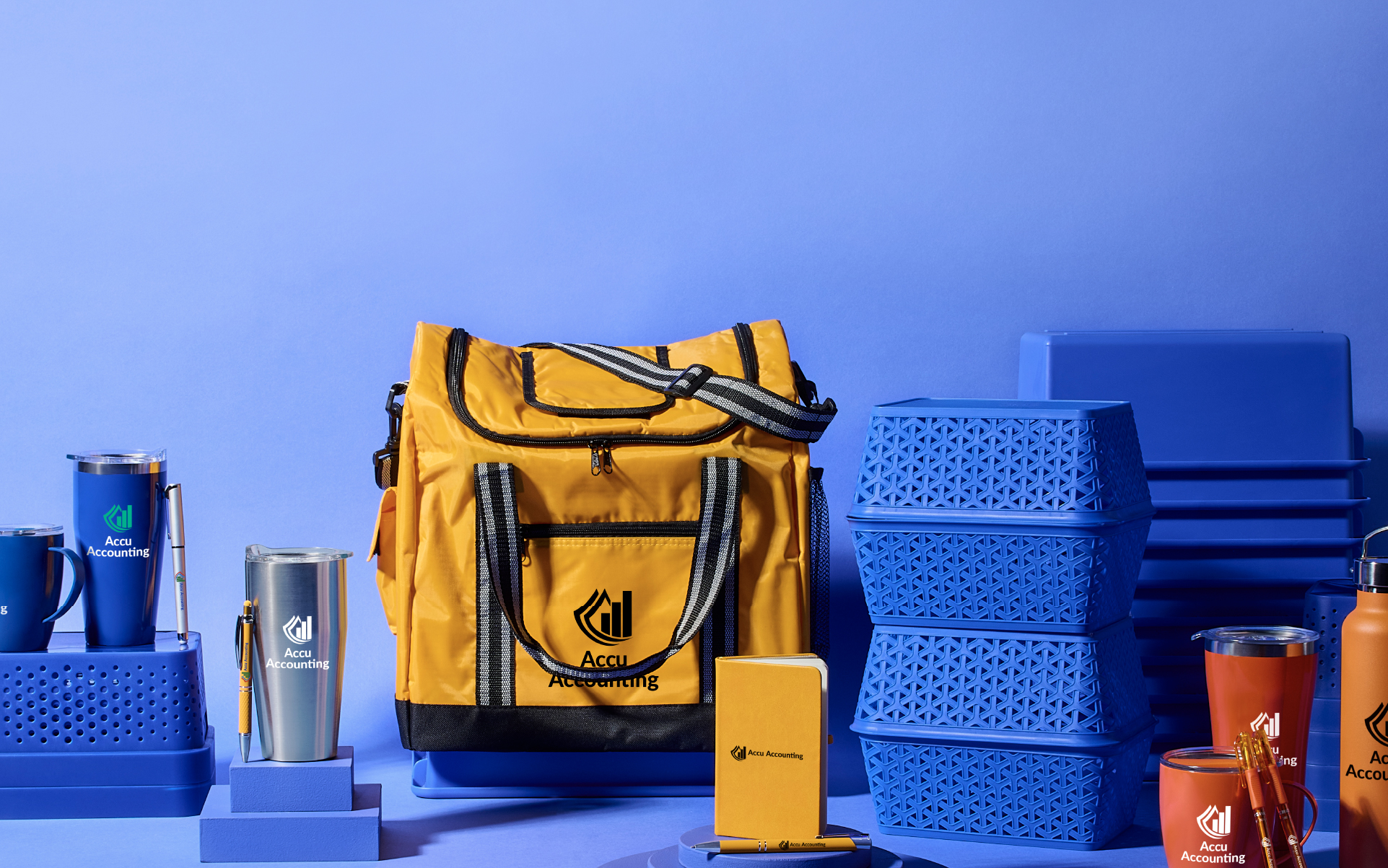 A variety of trade show promotional products on display against a blue background - metal tumblers with logos, orange bags, notebooks, lanyards and water bottles.