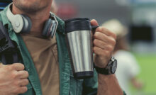 Mensch im Freien mit Edelstahl-Thermobecher befüllt mit Heißgetränk in der Hand.